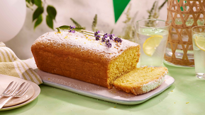 A golden lemon drizzle loaf cake topped with powdered sugar and sprigs of fresh lavender, resting on a white rectangular plate. A slice has been cut to reveal its soft, fluffy interior.