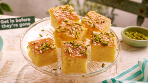Ravani sponge cake sprinkled with pistachios, cut into squares and displayed on a cake stand.