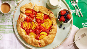 A golden-brown rustic fruit galette brimming with juicy peach and strawberry slices sits on a light blue wooden board. It's surrounded by fresh berries, steaming cups of tea or coffee, and a pitcher of cream.