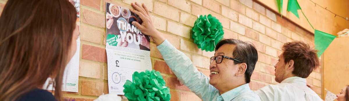 A group of people are putting up decorations on a brick wall for a Coffee Morning. One person in a light blue shirt with short black hair is holding up a poster to the wall. They appear to be in an office. 
