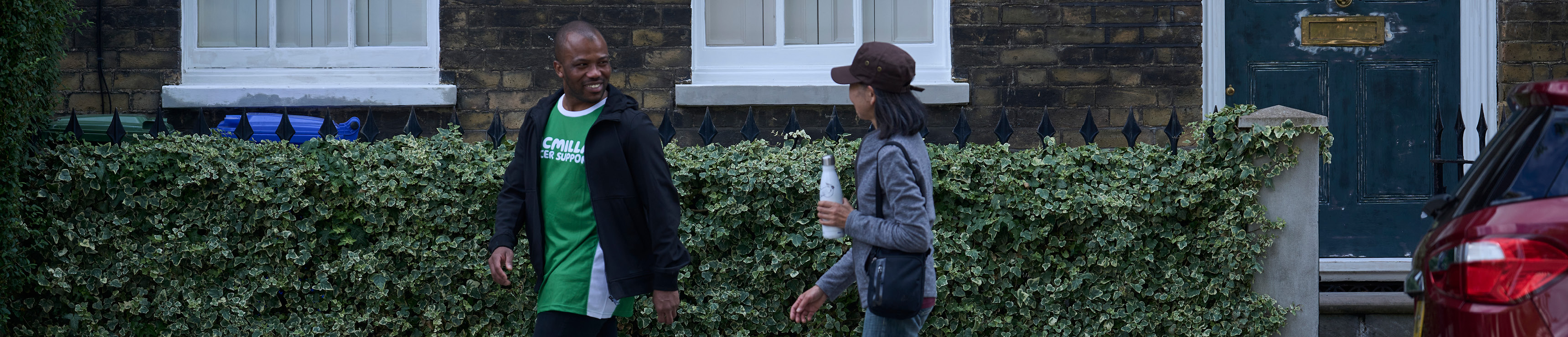A man wearing a Macmillan t-shirt turning to talk to someone on the street outside a house 