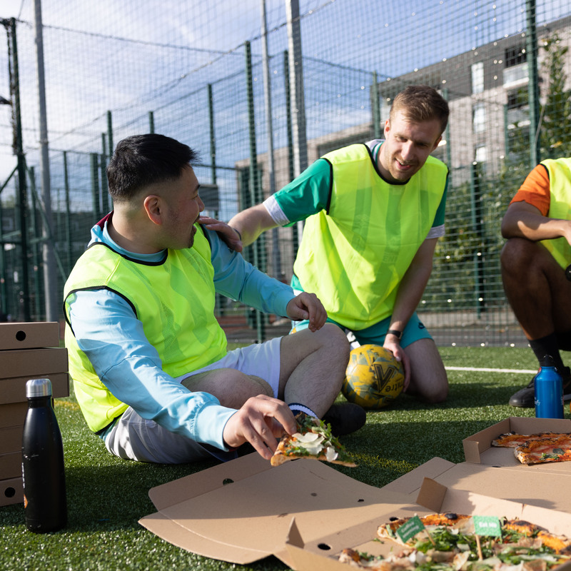 Three people are sitting down outside on a football pitch. They are all wearing football gear, including bright vest tops. They are boxes of pizzas around them, with one person holding a slice from an open box. 