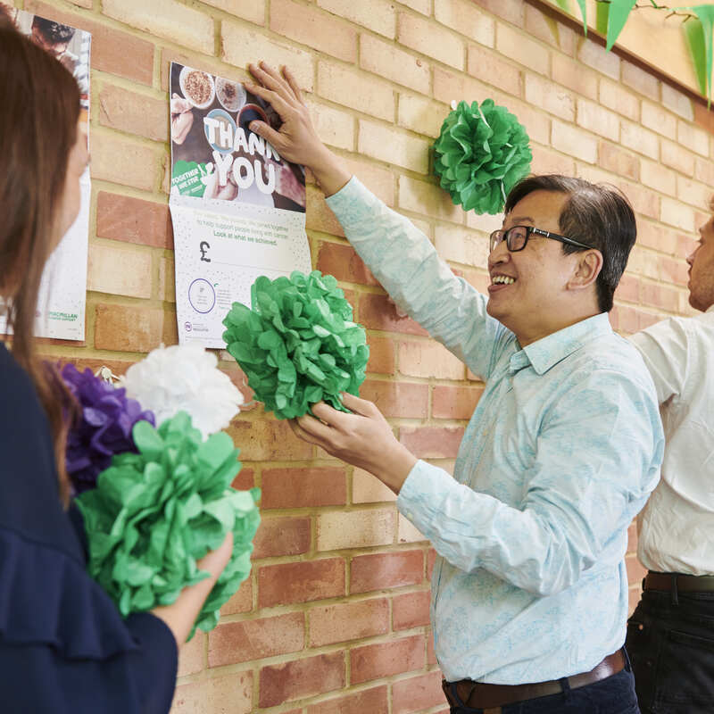 A group of people are putting up decorations on a brick wall for a Coffee Morning. One person in a light blue shirt with short black hair is holding up a poster to the wall. They appear to be in an office. 