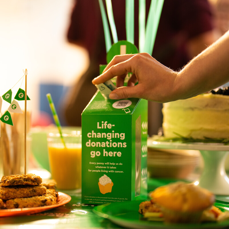 A person is putting a £5 note in a cardboard Macmillan branded donation box. The box is on a table with many cakes and other treats. 