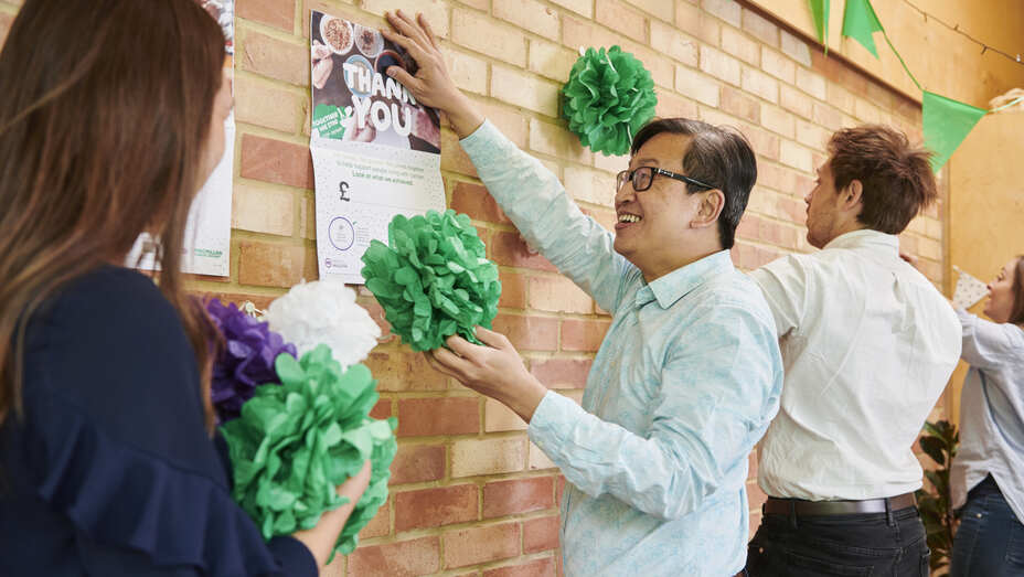 A group of people are putting up decorations on a brick wall for a Coffee Morning. One person in a light blue shirt with short black hair is holding up a poster to the wall. They appear to be in an office. 