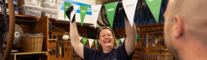 A person in a dark blue top has both arms in the air as they try to hang Macmillan branded bunting. They have short blonde hair and are also wearing black gloves. Behind them are different tools and equipment for bike repairs. 