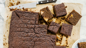 A pear and ginger cake is on brown baking parchment. Part of the cake has been cut into slices. Next to the cake is a silver knife. There is also a plate with some slices of the cake on it.