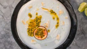 A view of a vegan lime, passionfruit and coconut cake from above. The cake is topped with a white frosting, half a passionfruit, and passionfruit seeds. It is in a dark rimmed container. 