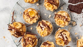 Several vegan peanut butter and banana cupcakes are on a cooling rack. The top of the cupcakes are decorated with frosting, chocolate flakes, and baked banana pieces. Next to the cupcakes is a broken bar of chocolate. 
