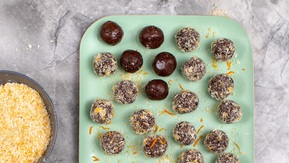 Several truffles are placed on a mint green plate. The truffles are covered in chocolate, and some have a sprinkle of desiccated coconut. Next to the plate is a bowl of desiccated coconut.