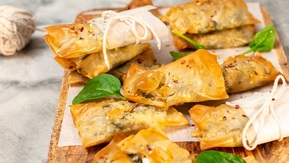 Several spinach and feta parcels are on a wooden board. Some of the parcels have baking parchment and string wrapped around them. Also scattered on the board are spinach leaves. 