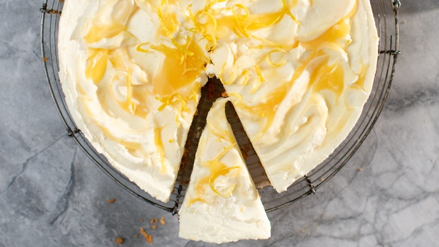 A creamy lemon cheesecake with swirls of glossy lemon curd and delicate strips of lemon zest sits on a wire rack. One slice is cut and slightly pulled out, revealing its rich, velvety interior.