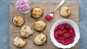 Seven golden-brown coconut macaroons with toasted edges rest on a wooden board, paired with a bowl of vibrant raspberry jam and a spoonful ready to serve.