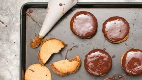A baking tray filled with round, golden cookies—some topped with glossy chocolate. A piping bag oozes rich caramel in the corner, white crumbled cookie pieces reveal a soft, crumbly texture.