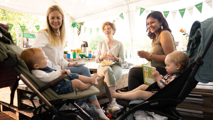 Three women are sitting down together. Next to two of the women are two strollers with babies in them. Behind the group are tables with food and decorations. 