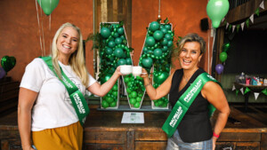 Two people are standing and smiling at the camera whilst clinking their white coffee cups together. The person on the left has long blonde hair and is wearing a white top and orange trousers. The person on the right has short hair and is wearing a black vest and blue jeans. Behind them is a balloon arch shaped like the letter M. 