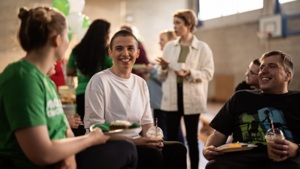 Three people are sitting and chatting at a Coffee Morning. A person with short hair and wearing a white tshirt is holding a plastic drink cup. Next to them is a person in a black tshirt holding a plate with food and a plastic drink up. Behind them are other people at the event. They appear to be in an outdoor basketball court. 