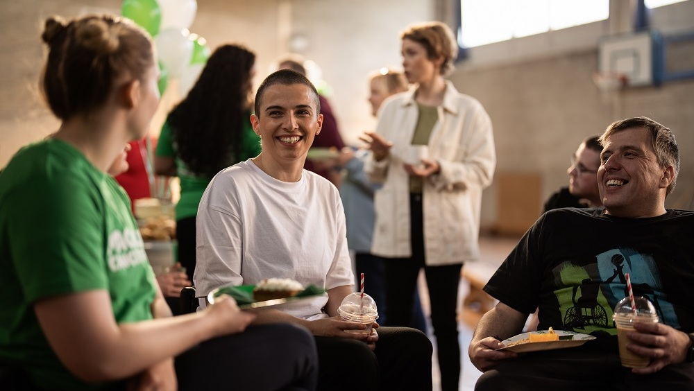 Three people are sitting and chatting at a Coffee Morning. A person with short hair and wearing a white tshirt is holding a plastic drink cup. Next to them is a person in a black tshirt holding a plate with food and a plastic drink up. Behind them are other people at the event. They appear to be in an outdoor basketball court. 