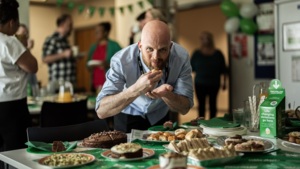 A man is leaning over a Coffee Morning table and taking a bite from a pastry. The man has on a blue shirt. The table has many different desserts and other treats on it. Behind the man are other people eating food and socialising. 