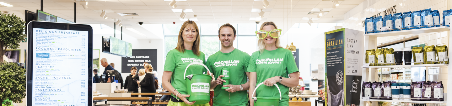 Three volunteers in green Macmillan branded tops are standing together in front of an M&S Café. Two of the volunteers are holding Macmillan branded collection buckets. A volunteer in the middle is holding a small Macmillan branded collection tin. 