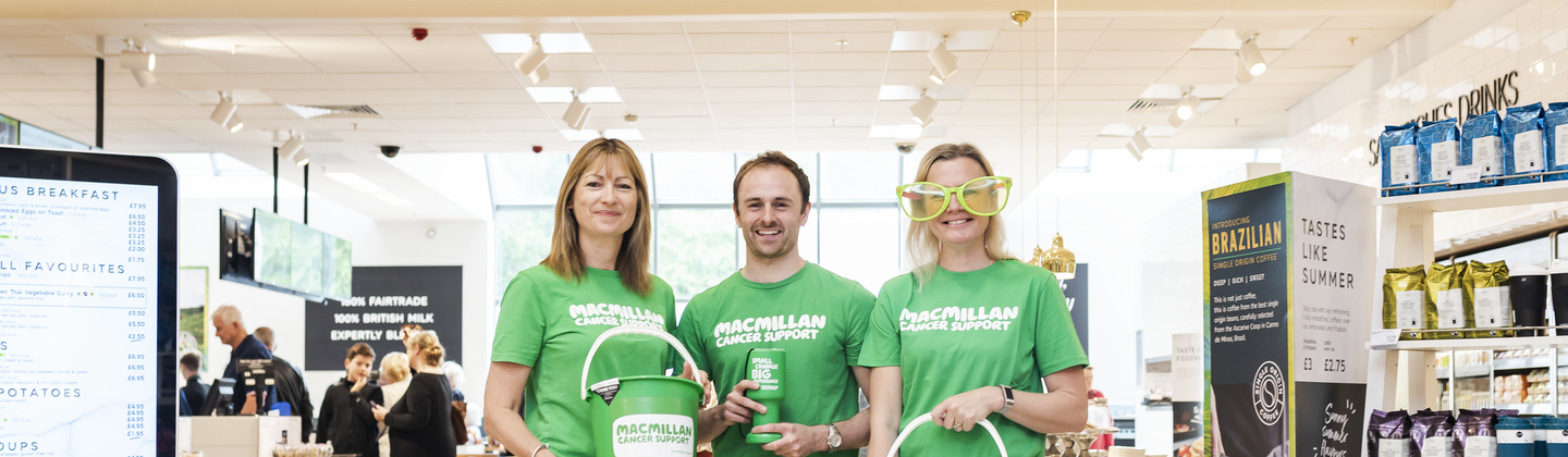 Three volunteers in green Macmillan branded tops are standing together in front of an M&S Café. Two of the volunteers are holding Macmillan branded collection buckets. A volunteer in the middle is holding a small Macmillan branded collection tin. 