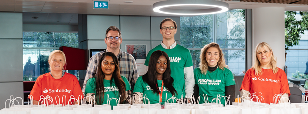 A group of people in Santander and Macmillan t-shirts standing behind a table covered in white paper gift bags.