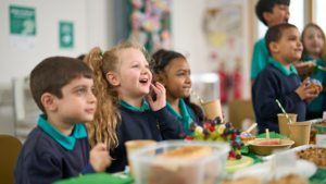 A group of kids are sitting at a table that has different treats on it. The kids are all in a dark blue uniform. They are looking at something not pictured in the photo.