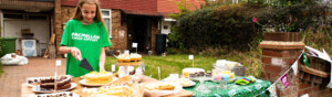 A young person is standing in a drive way. They have set up a cake selling booth on different tables. The young person is wearing a green Macmillan t-shirt. They are cutting a slice of cake. The tables have different cakes and other treats for sale. 