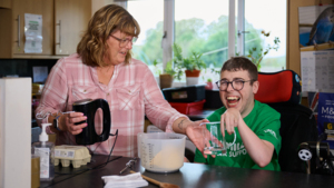 A woman with short hair wearing a pink chequered top is standing in a kitchen near a counter and holding an electric mixer. Next to hear is a man sitting in a wheel chair and wearing a green Macmillan top. He is passing the woman a glass container. They appear to be preparing ingredients for baking.
