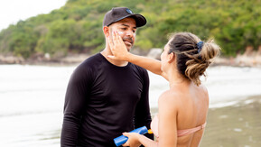 Woman applying sunscreen to mans face while at a beach.