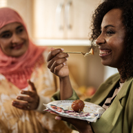 Two women in a kitchen eating cake.