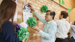 A group of people are putting up decorations on a brick wall for a Coffee Morning. One person in a light blue shirt with short black hair is holding up a poster to the wall. They appear to be in an office. 