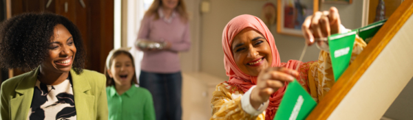 A person in a pink headscarf is placing Macmillan branded bunting up in a hallway in their home. To their left is a person holding a tin foil wrapped dish. There is also other people in the photo including a young person in a green outfit and an older person wearing a denim skit and pink top. 