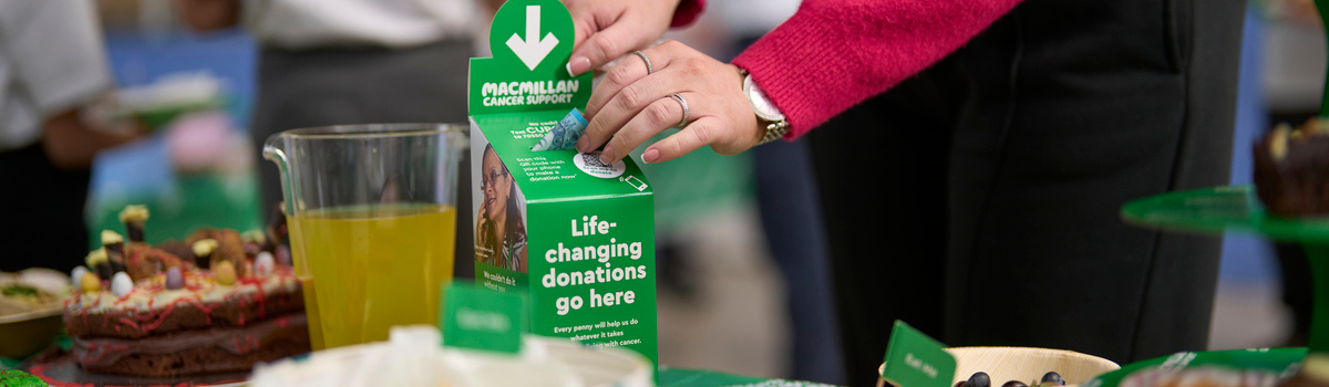 A person in a bright pink jumper is leaning over a table and placing a pound note in a Macmillan collection box. The box is on a table that has been decorated for a Coffee Morning. There are drinks and different sweat and savoury foods on the table. 