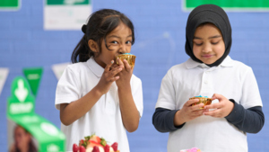 Two young people in school uniforms are holding cupcakes. The person on the left is biting into the cupcake. They appear to be in a school. 