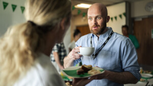A person in a blue shirt and short, brown beard is holding a large white coffee cup in their hand. They are speaking to a person with long blonde hair wearing a white shirt. The person in the white shirt is holding a plate with food on it. They appear to be inside.