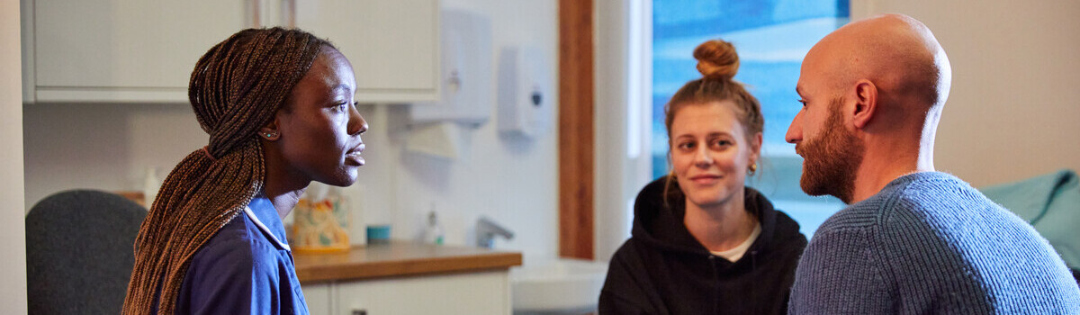 Pictured left is a nurse who is sat on a chair. She is facing a man who is wearing a blue jumper. Further away a woman in a black hoodie is listening to the nurse speak to the man.