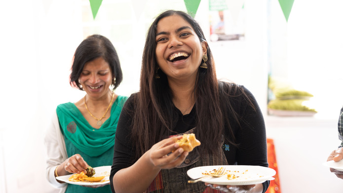 A person with long dark hair is laughing. They are holding a treat in one hand and a plate with a fork in the other. Behind them is another person with short hair who is also laughing and holding food and a plate. They appear to be inside. 