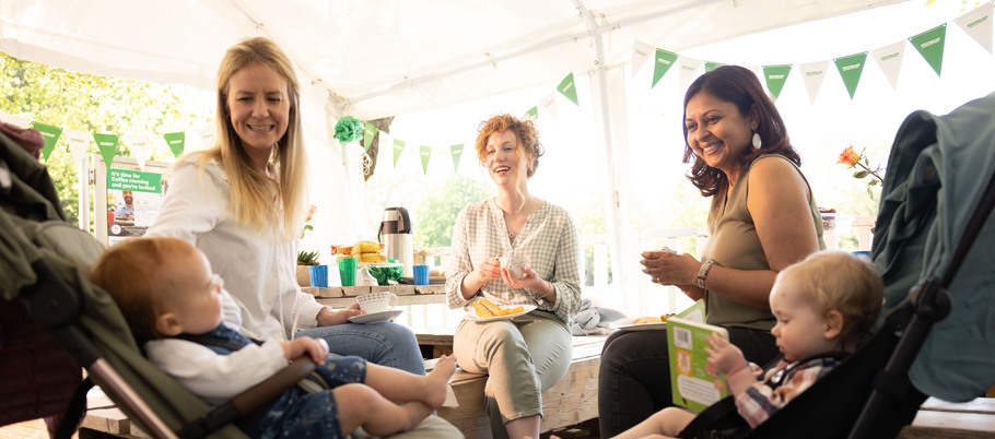 Three women are sitting down together. Next to two of the women are two strollers with babies in them. Behind the group are tables with food and decorations. 
