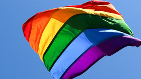 Two rainbow flags being waved against the backdrop of a blue sky.