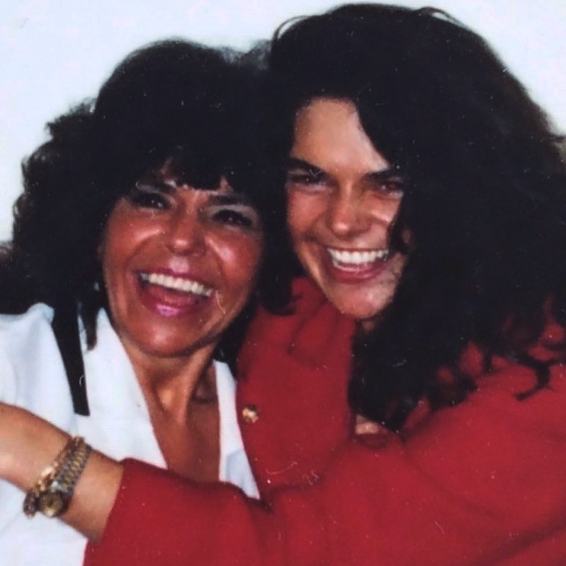 Two women with dark hair smiling and hugging. One is wearing white and the other is wearing red
