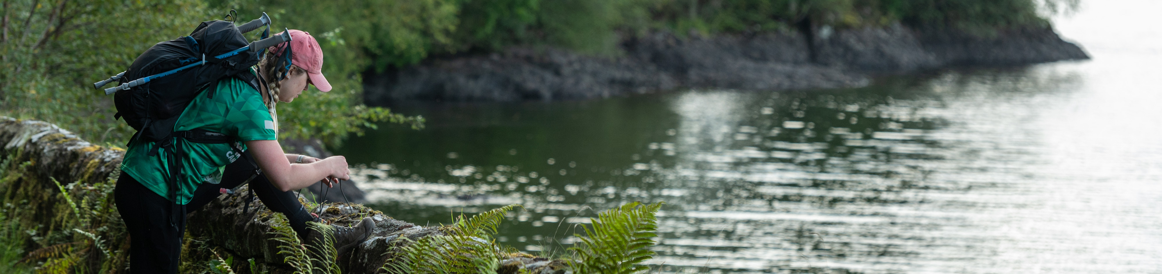 A Macmillan hiker by a lake doing up their bootlace on a wall