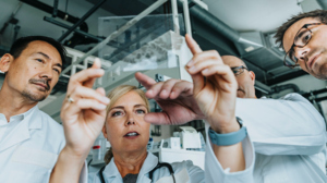 Four individuals are inspecting a glass that has a human sample it in. The person in the middle is holding up the glass in front of the others. The person to their right is pointing at something in the sample. 