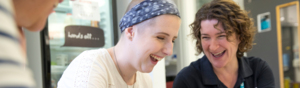Jamie Cargill (right) is with a young patient (middle). They are both laughing. The young patient is holding a tablet in her hands. They are sat at a table indoors.