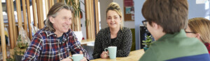 A group of people are sitting at a table at a Macmillan support centre. Some of the people in the image are holding coffee mugs in their hands.