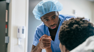 A nurse in blue scrubs and wearing a blue scrub cap is holding the hand of a patient who is laying down in a medical bed.