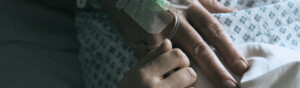 A hand with a cannula in it is resting on a patients body. There is another hand that is holding a finger of the patient with the cannula.