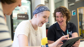 Jamie Cargill (right) is with a young patient (middle). They are both laughing. The young patient is holding a tablet in her hands. They are sat at a table indoors.