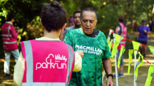 A person in a green Macmillan branded running top is being handed something by a person in a pink parkrun branded vest. They appear to be outside at a running event.