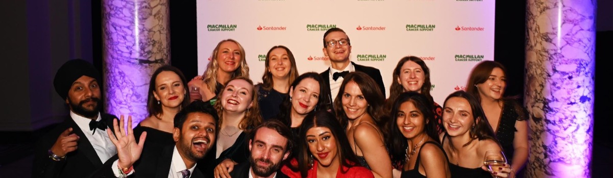 A large group of people in formal wear are standing in a group posing for a photo. They appear to be indoors and are standing in front of a white banner that has Macmillan and Santandar logos on it.